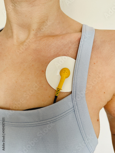 Close-up of a female chest with a yellow ECG electrode attached to the skin for medical heart monitoring purposes. Shot indoors against neutral background.