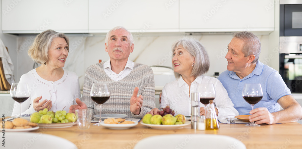 Four mature male and female friends are sitting at table, chatting sweetly, discussing and telling news, sharing plans. Senior women and men celebrates housewarming party with female relative. Four mature male and female friends are sitting at table, chatting sweetly, discussing and telling news, sharing plans. Senior women and men celebrates housewarming party with female relative.