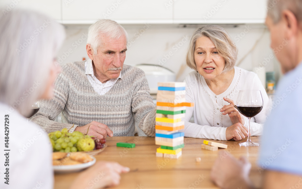 Elderly happy people playing Jenga at home, woman is removing a wooden block from the tower and the others are watching Elderly happy people playing Jenga at home, woman is removing a wooden block from the tower and the others are watching