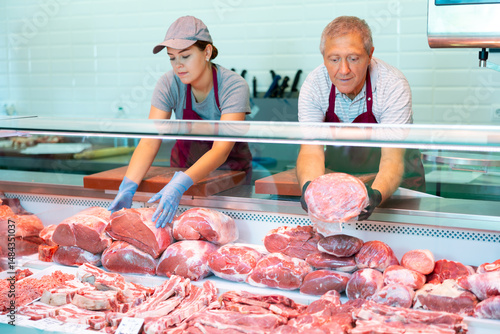 Focused skilled workers of butchery shop, elderly man and young woman, working together behind counter, arranging fresh raw meat cuts in glass refrigerated showcase