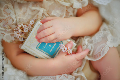 Hands of a newborn baby holding a small book and dry flowers. Baby in a vintage dress. The girl is 5 days old. Just born. Small fingers of a newborn baby. Flowers in hands. Book in hands. Portrait.
