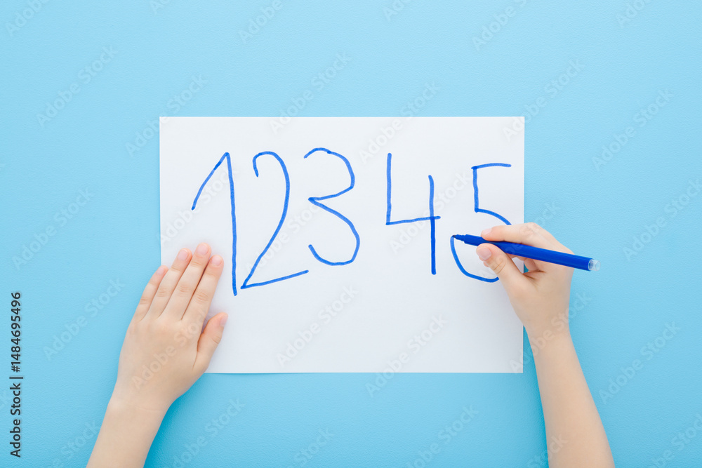 Fototapeta premium Little child boy hand holding color pen and writing different numbers on white paper on light blue table background. Pastel color. Doing school exercise. Closeup. Point of view shot. Top down view.