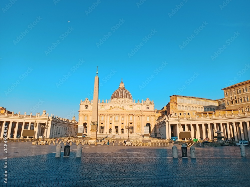 Obraz premium Stunning view of St. Peter’s Basilica with the Vatican Obelisk under a clear blue sky at sunrise. A symbol of Rome’s architectural and religious grandeur.