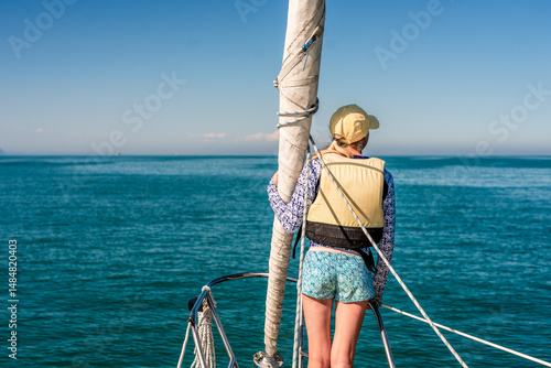Active girl in yellow hat on bow of sailboat looking out over ocean