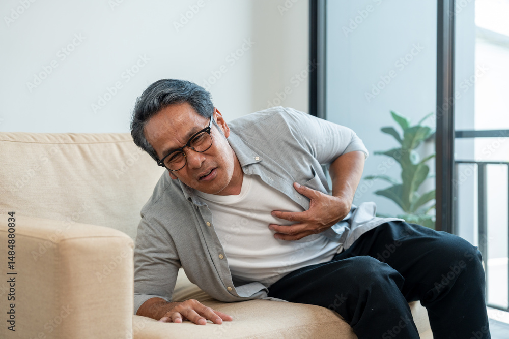 Fototapeta premium Old man senior he felt a pain in his chest held his hand and squeezed it. Mature Heart attack that needed treatment. Sitting on sofa couch in living room at house