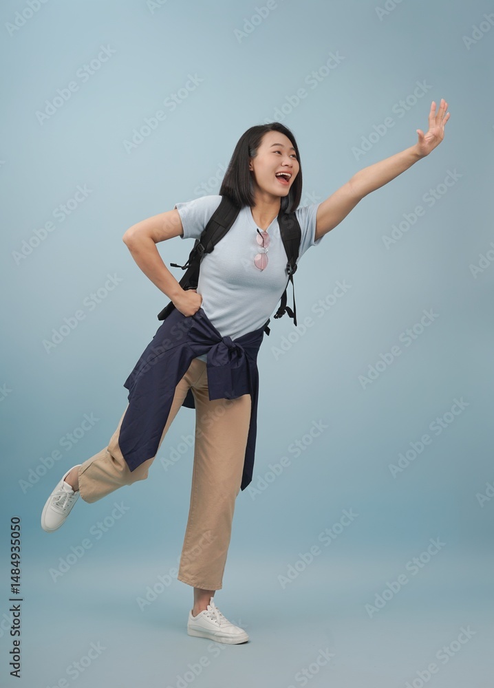 Obraz premium Happy young woman joyfully posing with one leg raised against a light blue background in a casual outfit and backpack