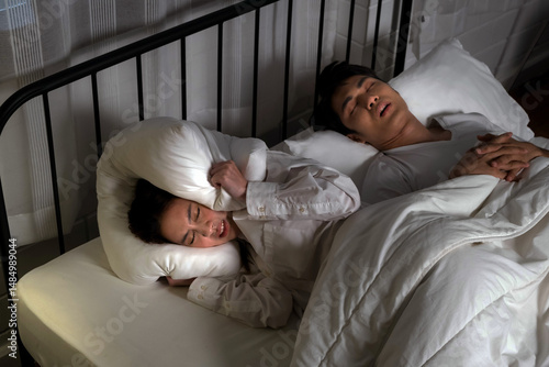 Woman struggles to sleep beside snoring partner, covering ears with pillow in dark bedroom. Concept of relationship tension, sleep disturbance, and untreated obstructive sleep apnea (OSA).
