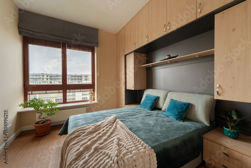 Bedroom with bed in alcove of built-in light wood cabinets. Modern room has ample storage, large window with blinds, and wood floor. Bright with natural light, this interior offers functional living.