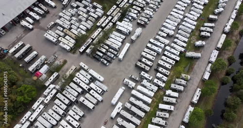 Large parking lot of local dealer with many new RV trailers parked for sale. Distribution of manufactured camping vehicles.
