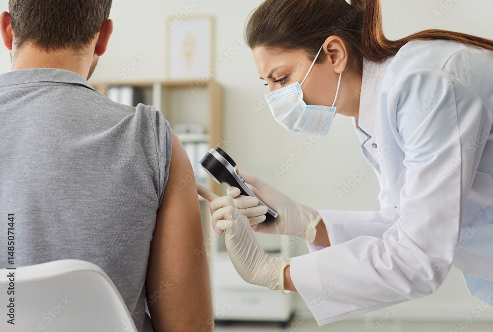 Fototapeta premium Female doctor examining hand skin of young man patient. Professional woman dermatologist using magnifying glass checking skin condition in clinic. Dermatology, skin health care concept.
