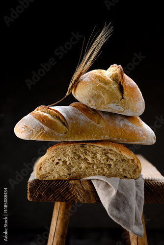 Composition from crusty sourdough baguette, bread loaf and bun on a rustic wooden stool with ear and linen towel. Artisan farmhouse-style loafs captured in warm natural lighting