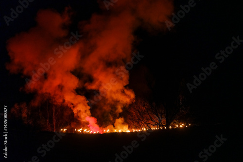 Krampus walk across a field in front of a wall of fire and smoke, silhouetted trees in the foreground. Krampus and St Nicholas, 5 December in Tarvisio, Friuli Venezia Giulia, Julian Alps, Italy.