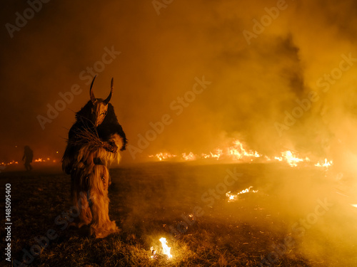 A Krampus figure in a furry suit and horned mask stands amidst smoke and fire. Another figure is visible in the background. Krampus and St Nicholas, 5 December in Tarvisio, Italy.
