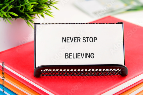 NEVER STOP BELIEVING motivational phrase on a white business card in a stand on a stack of notebooks