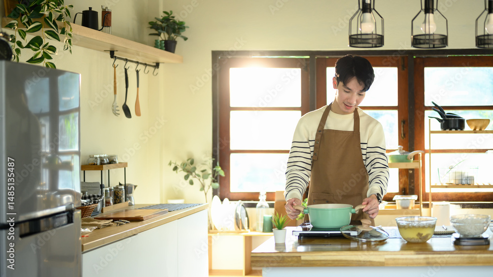 Young man smiling while preparing food in a home kitchen, standing over a pot on the stove Young man smiling while preparing food in a home kitchen, standing over a pot on the stove