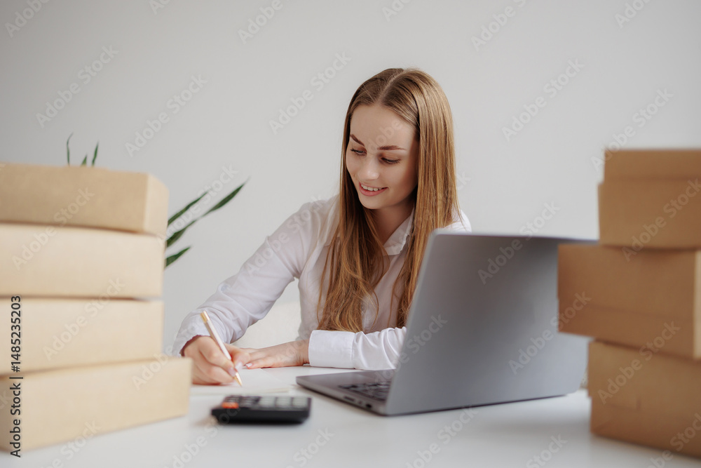 Young woman working at a desk surrounded by parcels and a laptop while taking notes in a bright workspace Young woman working at a desk surrounded by parcels and a laptop while taking notes in a bright workspace
