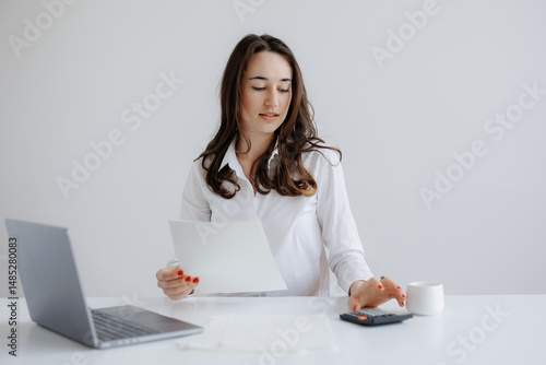 Young woman working on calculations with laptop, paper, and calculator in modern office setting