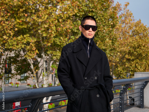Portrait of handsome Chinese young man wearing black overcoat posing in the street, young guy with black short hair all in black with urban background. Male fashion, cool Asian young man lifestyle.