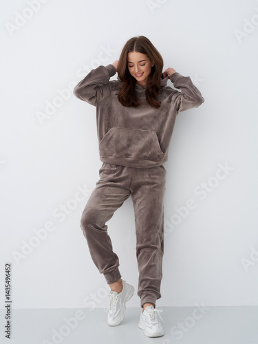 Woman in cozy brown loungewear set with a relaxed pose and white sneakers, adjusting her hair and smiling, standing against a simple white background. Comfortable, casual look