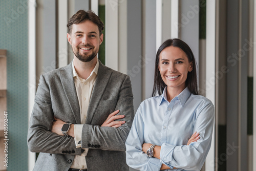 Portrait of smiling young Caucasian business man and business woman standing arms crossed in office looking at camera. Two diverse colleagues, group team of confident professional business people.