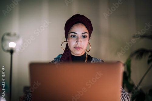 A young Moroccan Arab Muslim woman is focused on her laptop screen while working or studying indoors at night. She concentrates intently on the task.