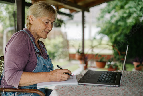 Elderly woman working from home. 
Concentrated good-looking senior woman writing notes in notepad while sitting at table in front of laptop in garden.