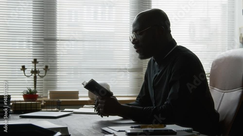 Side view of African American young priest with glasses wearing black shirt reading holy bible sitting at large table in contemplative manner