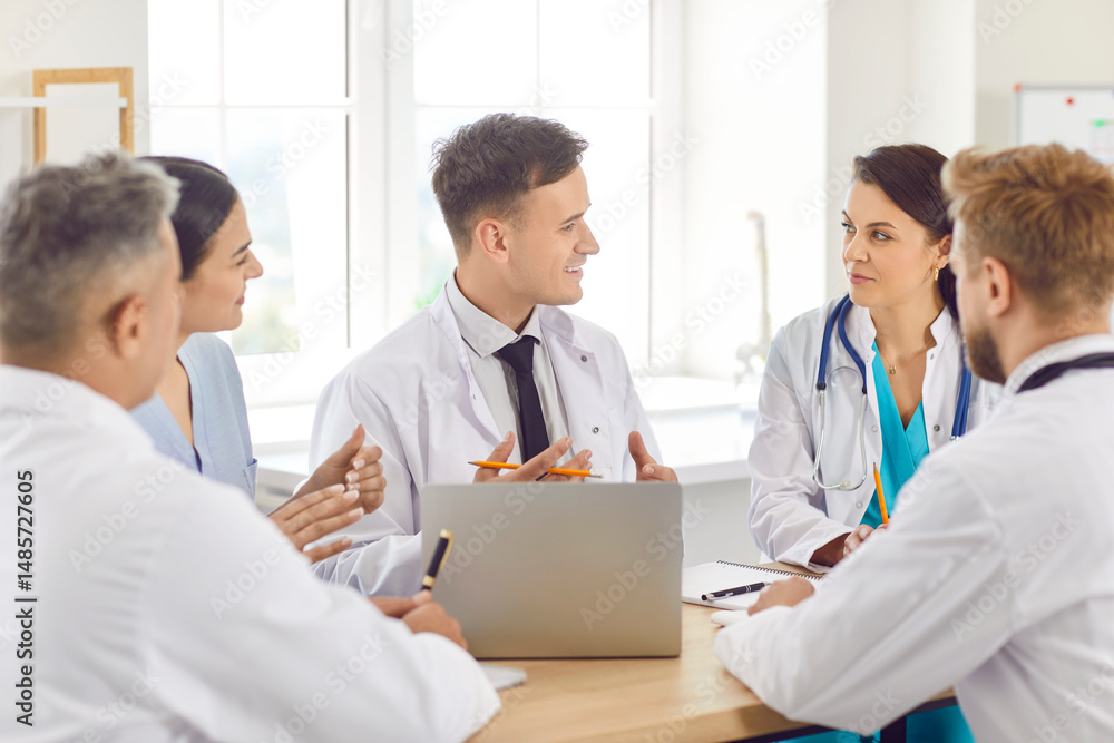 Happy young team of confident doctors in white uniform sitting on workplace in hospital or clinic on meeting discussing diagnosis or planning treatment for patients. Medicine and health care concept. Happy young team of confident doctors in white uniform sitting on workplace in hospital or clinic on meeting discussing diagnosis or planning treatment for patients. Medicine and health care concept.