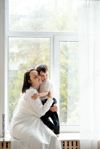 Free Download a mother with a small child on the windowsill, a mother with her son hugging and kissing, having fun together sitting on the window of the house, maternal love and care. - GFXLoad