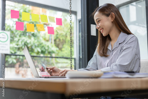 A female office worker is working at a desk with a laptop, credit cards, sticky notes and a pile of documents in front of her. She smiles, showing that she is happily working on a project.

