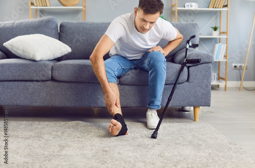Fotografie Unhealthy young man with foot trauma or sprain sitting on sofa next to the crutch, touching injured ankle with black elastic bandage and feeling pain