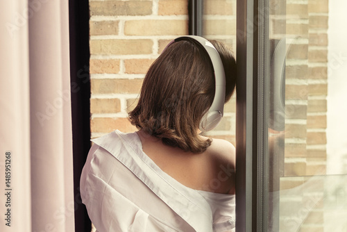 Back view of beautiful сaucasian woman in headphones enjoying listening music standing, leaning back on window.