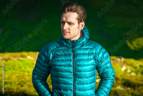 Portrait of a young man in a green down jacket looking away with serious face in windy mountain landscape