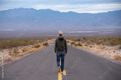 Man in a green sweatshirt and jeans walks alone on desert road toward mountains on Route 66 in Arizona.