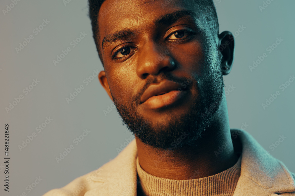 Obraz premium Confident young African man in stylish attire with soft lighting, showcasing a cool expression against a minimalist blue backdrop