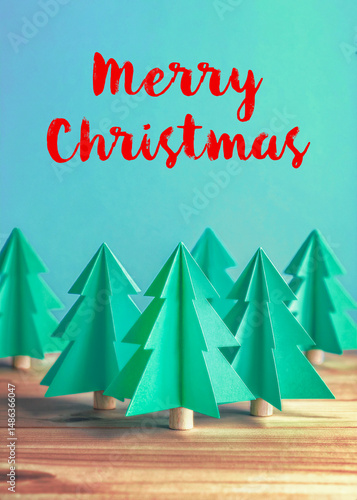 Trendy folded paper Christmas tree decorations on a wooden table against a turquoise green background that says Merry Christmas, easily replaced with your own personal message.