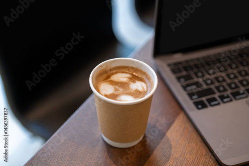 Close-up of Hot coffee latte with latte art milk foam in cup made of paper and laptop computer on wood desk office desk in coffee shop at the cafe,during business work concept