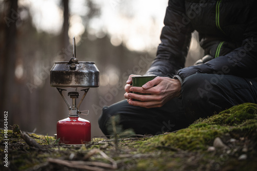 Person mit Tasse in der Hand vor Campingkocher in der Natur