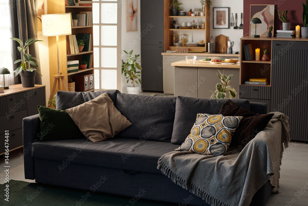 Cozy living room featuring a comfortable gray sofa adorned with patterned pillows and throw. Modern kitchen area visible in the background with some fresh fruits placed on counter Cozy living room featuring a comfortable gray sofa adorned with patterned pillows and throw. Modern kitchen area visible in the background with some fresh fruits placed on counter