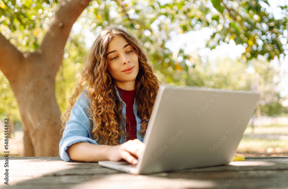 Stylish woman with laptop working at a wooden table in a sunny park. Beautiful female freelancer working outdoors. Remote work, leisure and education concept. Stylish woman with laptop working at a wooden table in a sunny park. Beautiful female freelancer working outdoors. Remote work, leisure and education concept.