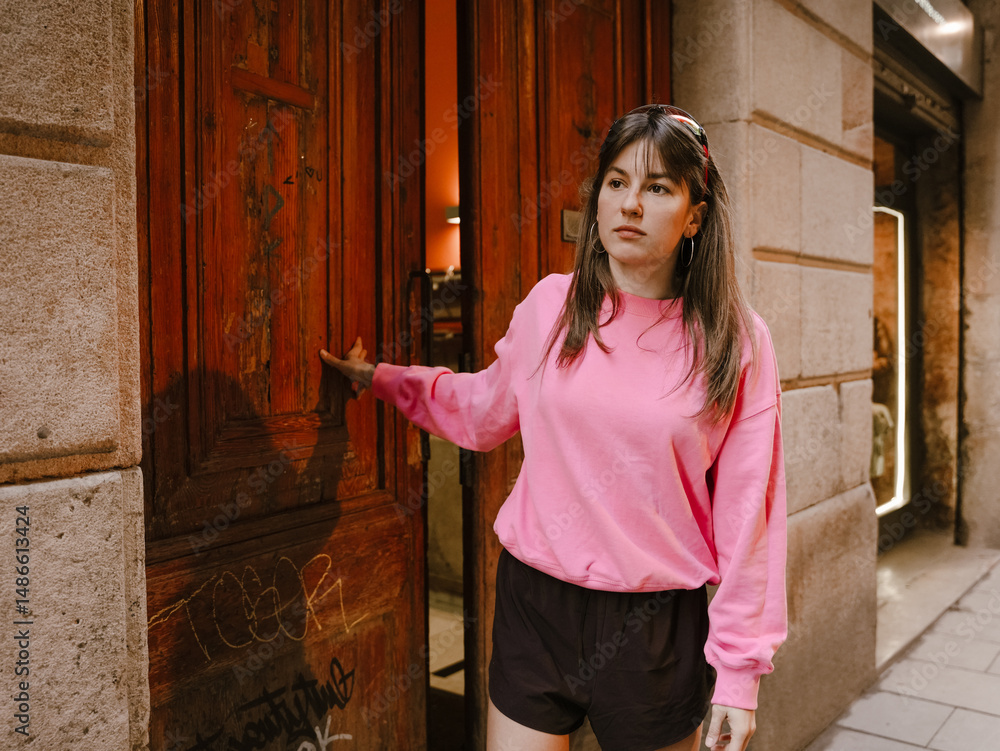 Fototapeta premium A young woman dressed in a bright pink sweatshirt and black shorts stands outdoors as she opens a large wooden door on a city street lined with stone walls and urban features.