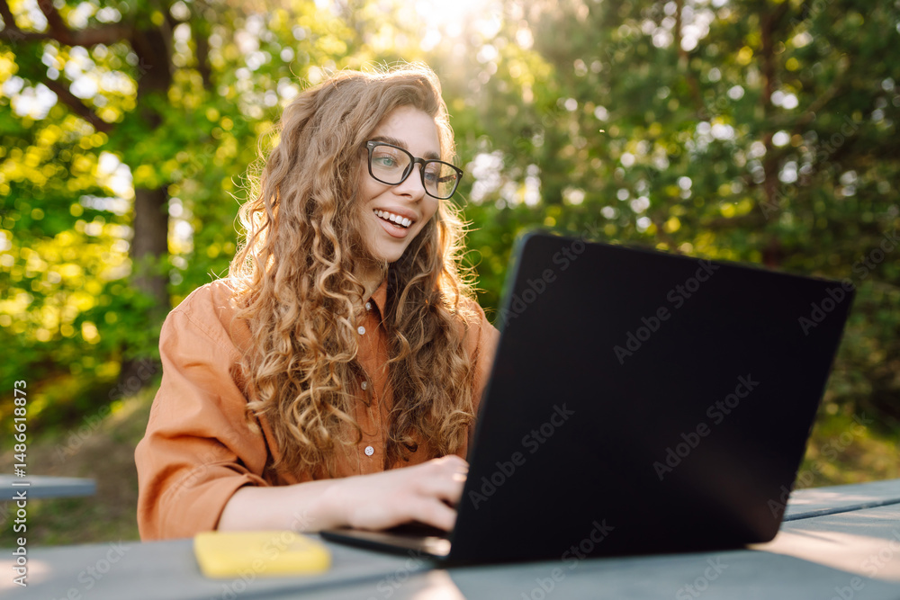 Portrait of young woman in glasses working on laptop in sunny park. Female freelancer at wooden table with laptop and phone outdoors. Remote work concept, technology. Portrait of young woman in glasses working on laptop in sunny park. Female freelancer at wooden table with laptop and phone outdoors. Remote work concept, technology.