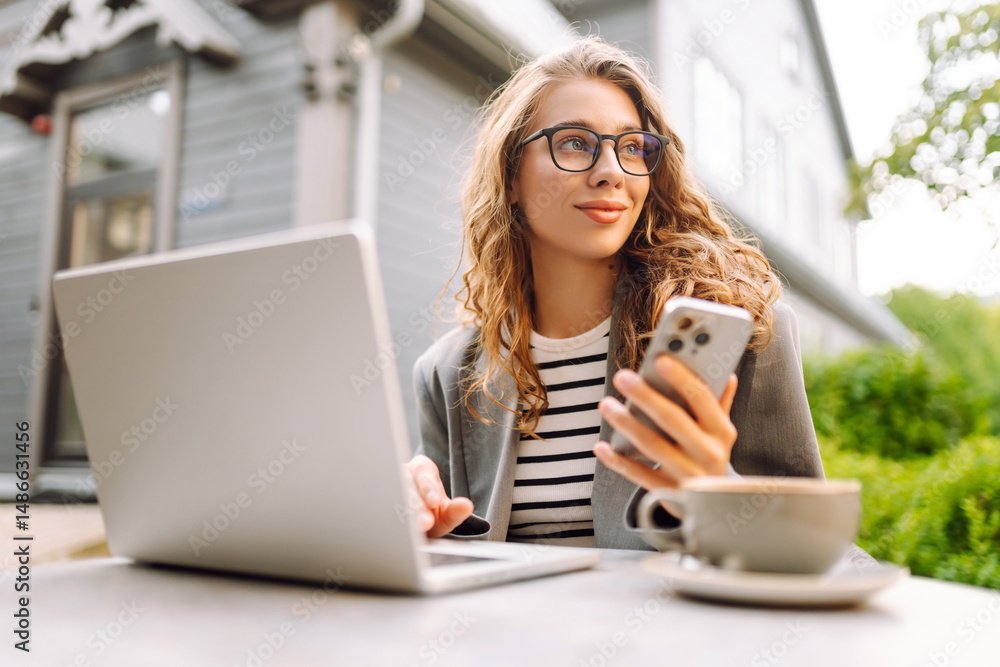 Woman in glasses works with a laptop on a summer terrace of a cafe with a phone in her hands. Female freelancer with phone and laptop enjoys the sunny weather. Freelancing, technology concept. Woman in glasses works with a laptop on a summer terrace of a cafe with a phone in her hands. Female freelancer with phone and laptop enjoys the sunny weather. Freelancing, technology concept.