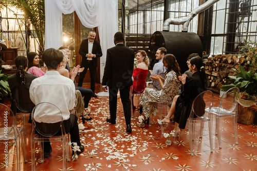 A dark-skinned groom in a suit is walking down an aisle covered with flower petals to a wedding arch and a beadle, while guests are sitting beside him, looking at him and clapping