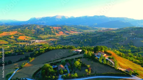 Detailed aerial push-in shot from San Ginesio, roads, buildings, trees and fields perched on suave hills touched by the warm afternoon sunlight, typical Marche landscapes and great Sibillini mountains
