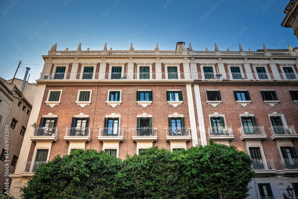 Naklejka premium Brick-and-stucco apartment façade in Ciutat Vella displays green-shuttered windows, iron balconies, neo-classical cornice above dense shrubbery, illustrating city-centre housing real estate heritage.