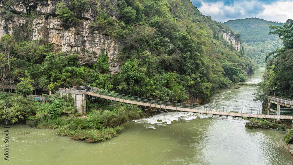Fototapeta premium A pedestrian suspension bridge spans over a mountain river. Water foams on boulders and rapids in the riverbed. Green vegetation on rocky slopes. Blue sky, clouds. China. Huangguoshu Waterfall.Guizhou