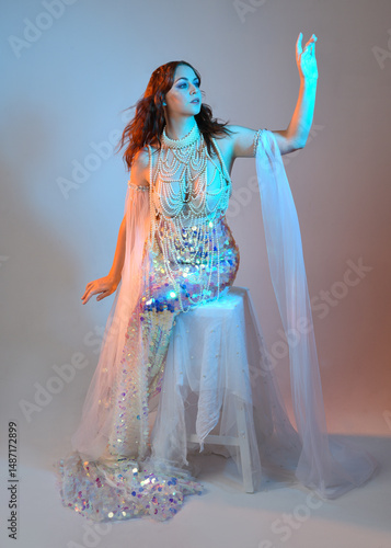 Fantasy studio portrait of a red-haired mermaid faery model wearing a white pearl gown, fairytale costume. classical sitting pose with flowing fabric and ethereal studio lighting, isolated background.
