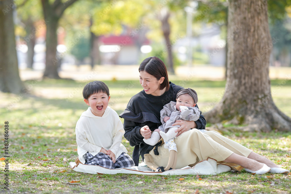 In spring at a park in Fukuoka, a mom in her thirties sits with her 3-year-old son and 6-month-old baby girl. They smile together. This photo shows a peaceful and heartwarming family moment in a sprin In spring at a park in Fukuoka, a mom in her thirties sits with her 3-year-old son and 6-month-old baby girl. They smile together. This photo shows a peaceful and heartwarming family moment in a sprin