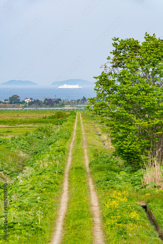 Obraz premium 春の田舎の景色 海の見える一本道の農道とフェリー 日本、青森